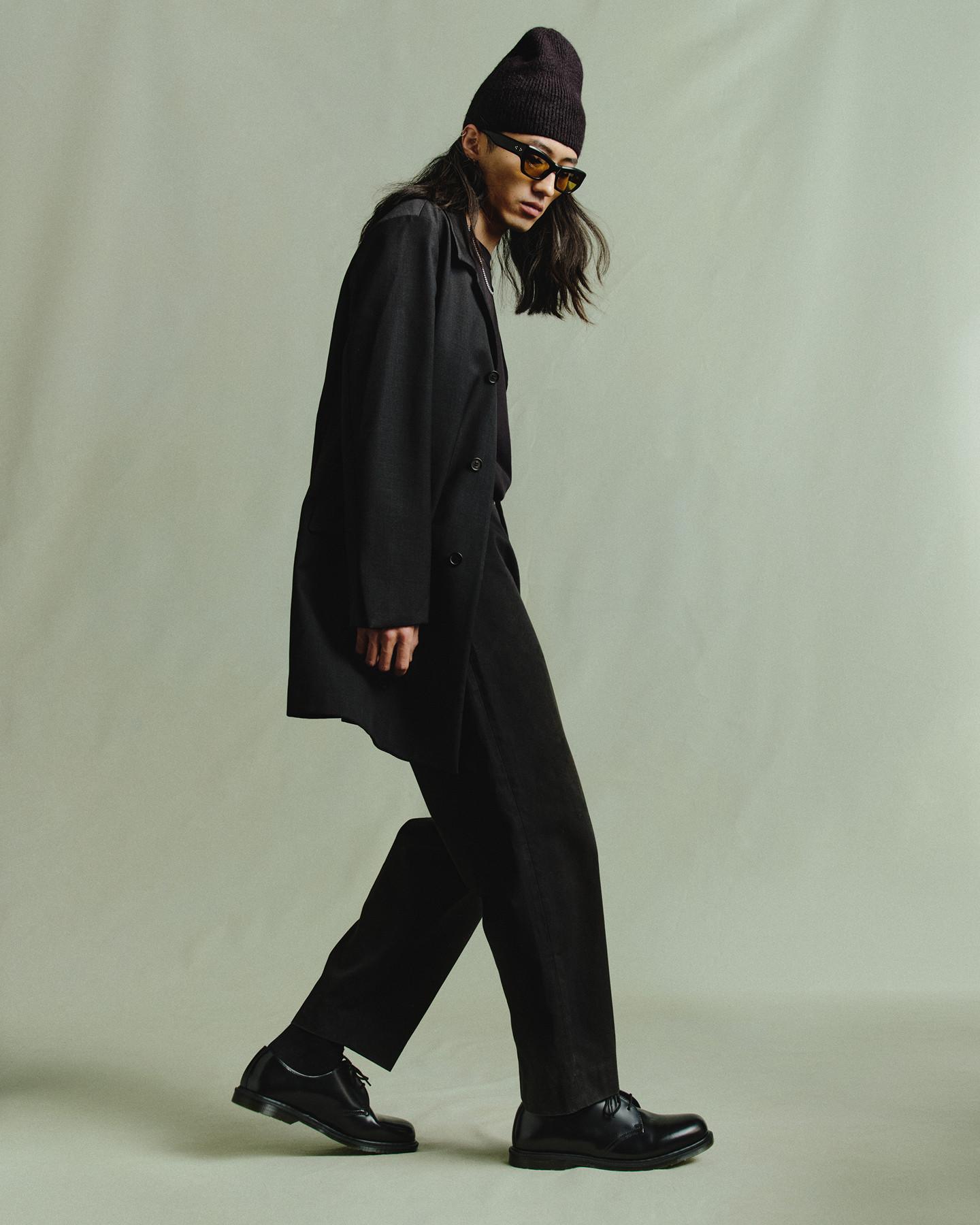 Persona con traje negro oversize, gorro de punto y gafas tintadas, caminando con zapatos negros