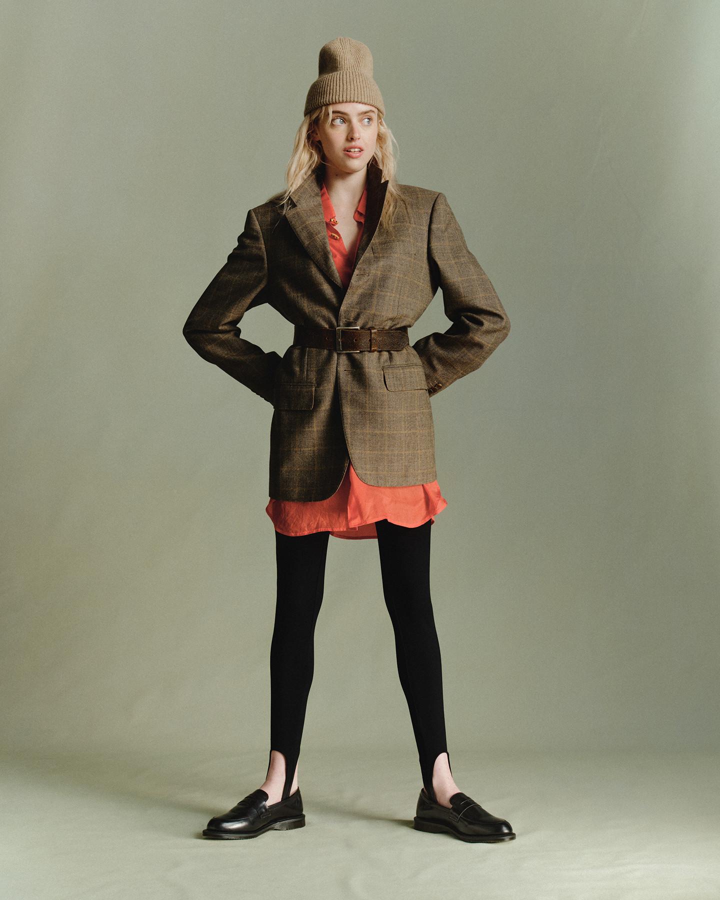Persona con mocasines negros de piel, medias y blazer de cuadros con cinturón sobre vestido camisero coral