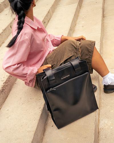 Black leather tote bag with wide straps on stone steps beside a seated person