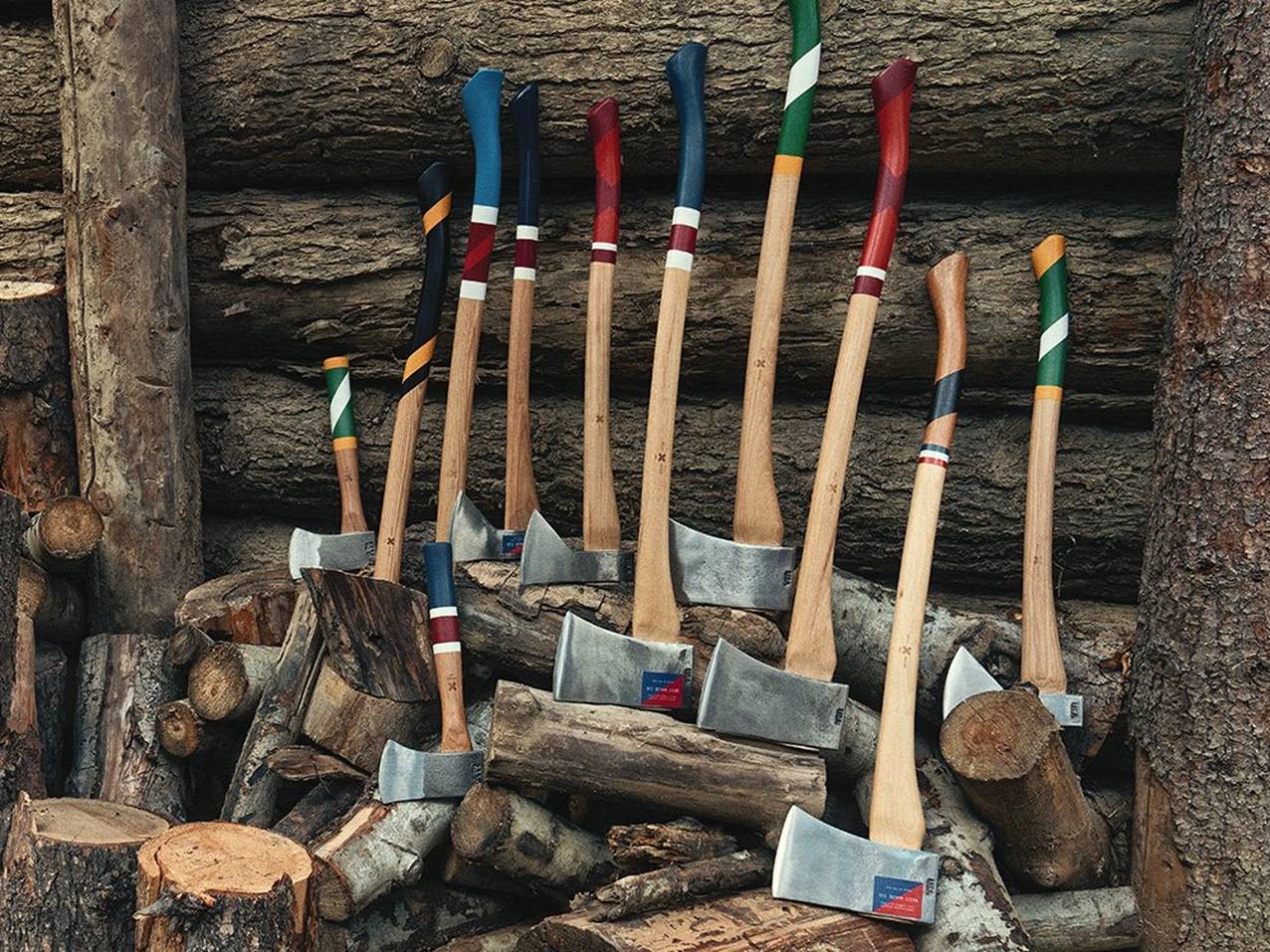 A collection of Best Made axes and hatches lean on a pile of cut wood against a log cabin