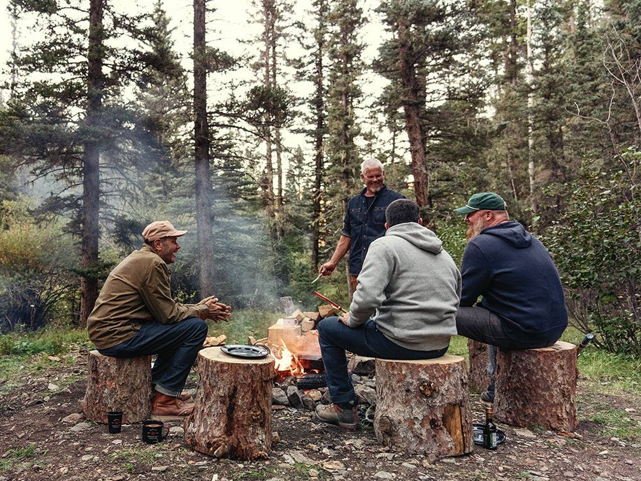 A group of man sit companionably around a campfire on wood logs.