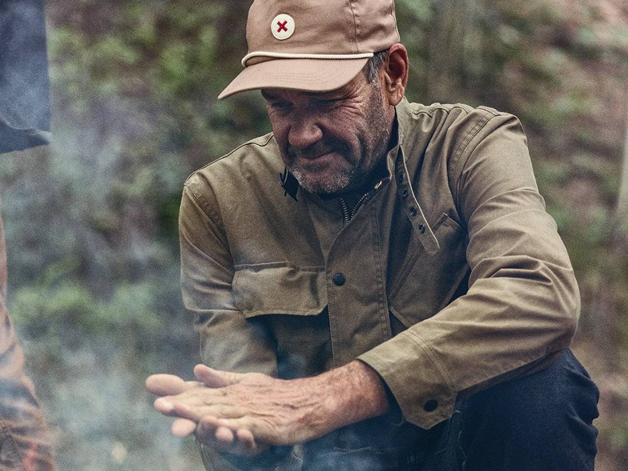 A man in a Best Made hat and khaki jacket sits at a camp fire.