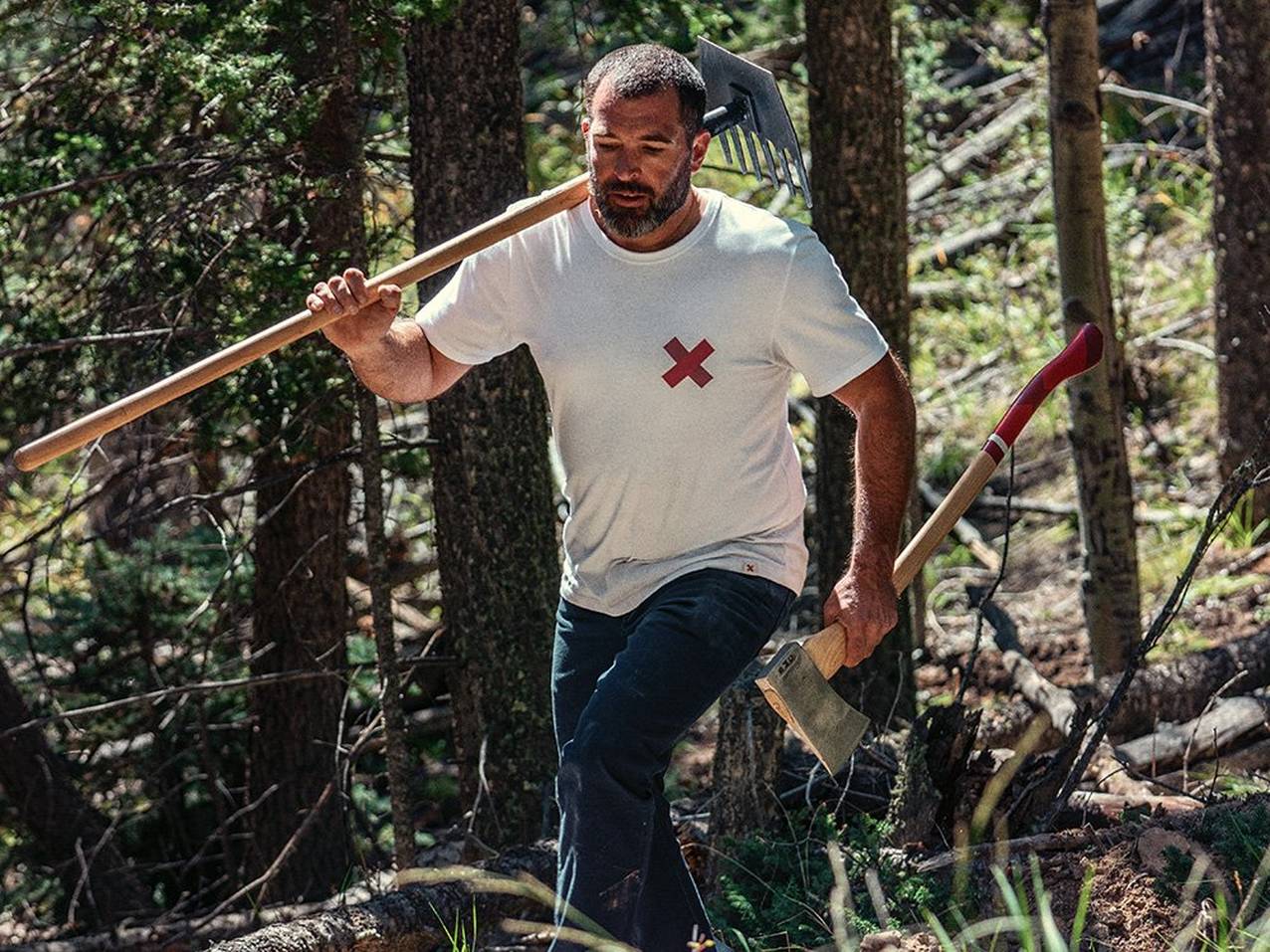A man in a white t-shirt with a red x on the left chest area walks up a hill in the woods carrying a rake and an axe