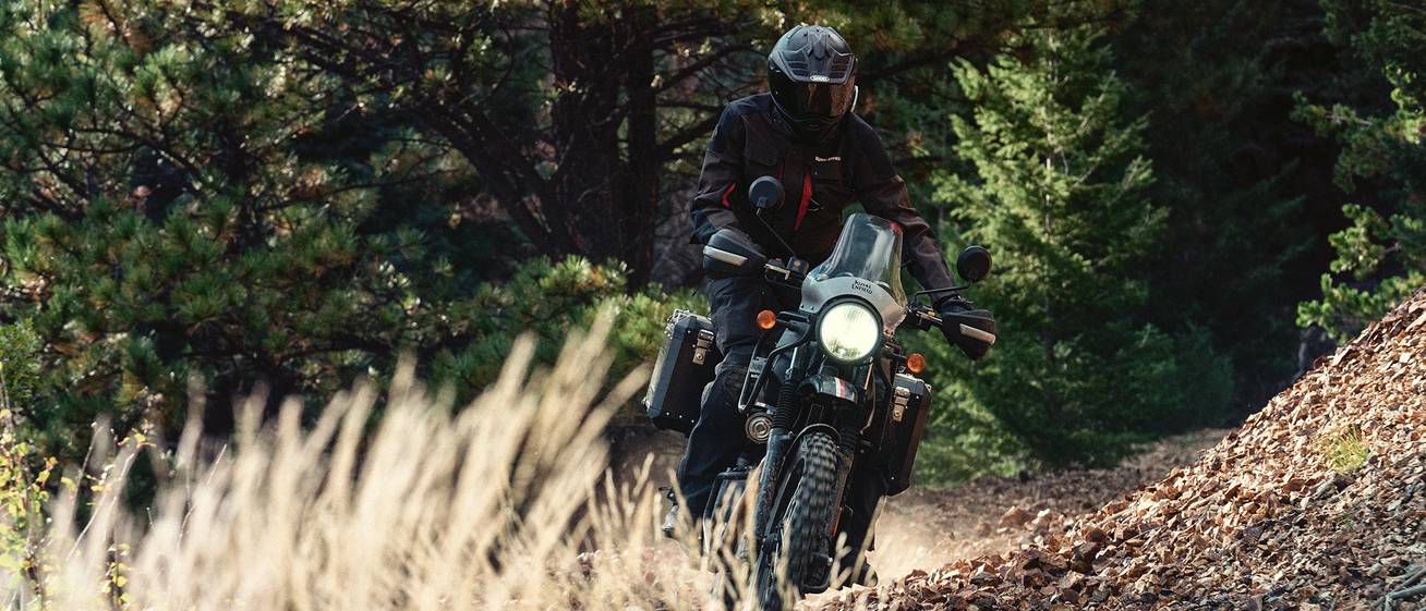 A man rides a motorcycle on a trail through the woods. His helmet and clothes are black with red accents.