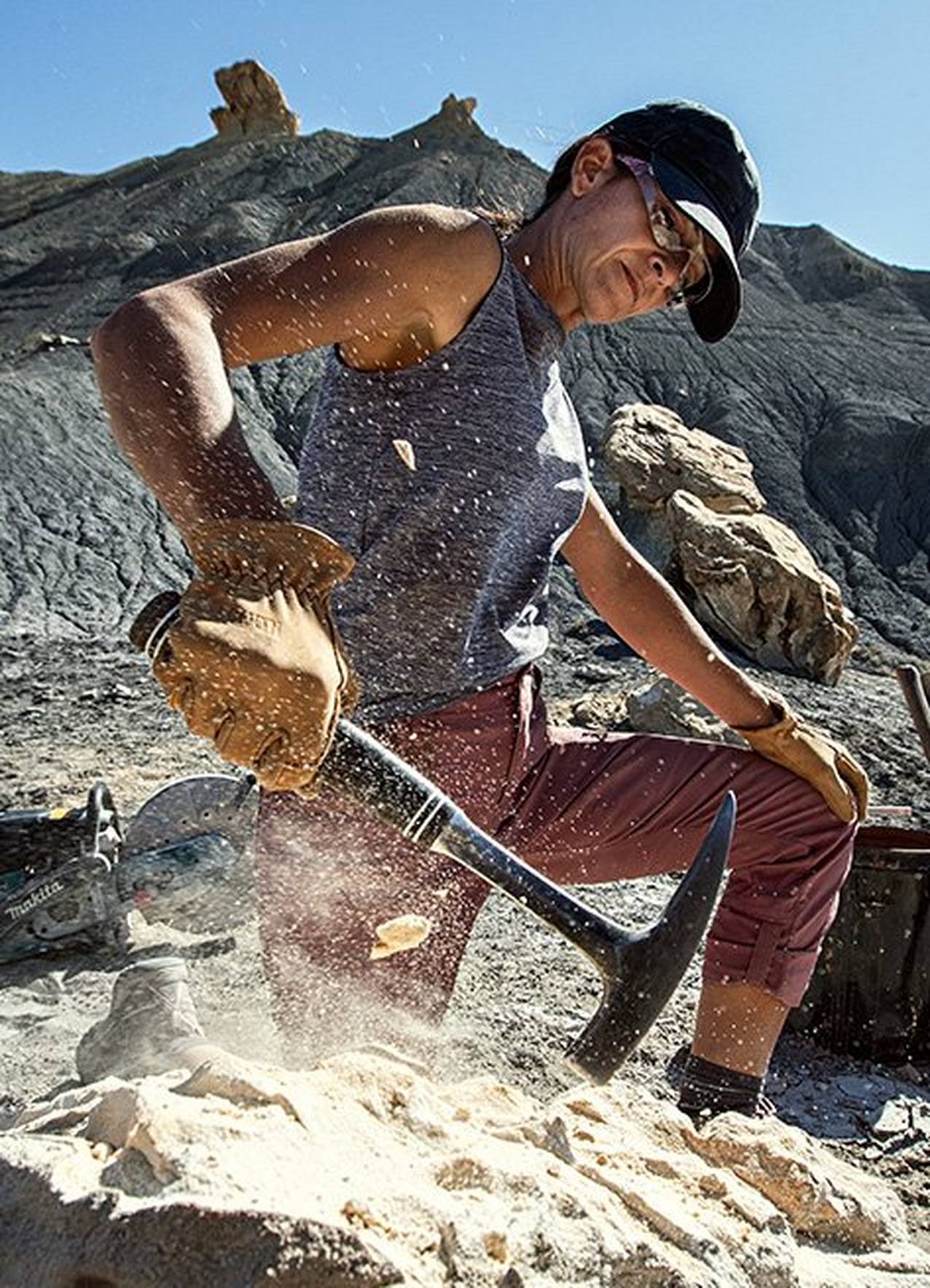 A woman uses a rock hammer in the hills of Utah. She is wearing pink dry on the fly capri pants and a gray tank top with protective gloves and glasses.