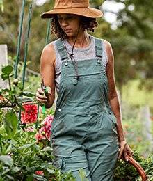 A woman in green overalls walks next to flowers holding a pruning shears