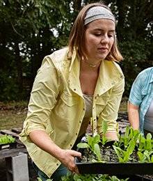 A woman in a yellow garden shirt holds a tray of seedlings