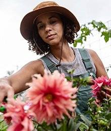 A woman in green heirloom garden overalls and a gray tank top leans forward to cut some pink flowers