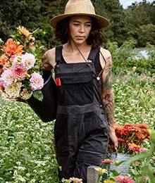 A woman in overalls walks with buckets of fresh-cut flowers