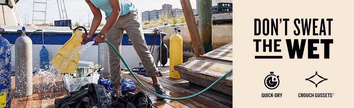 A woman hoses off her scuba gear on a pier wearing a blue tank top and khaki colored dry on the fly pants. Text that says don't sweat the wet on a cream background above two icons that indicate the technology of the product. One says quick-dry and the other crouch gussets(r)