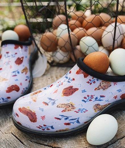 A pair of garden clogs with a chicken pattern sit next to a basket of eggs.
