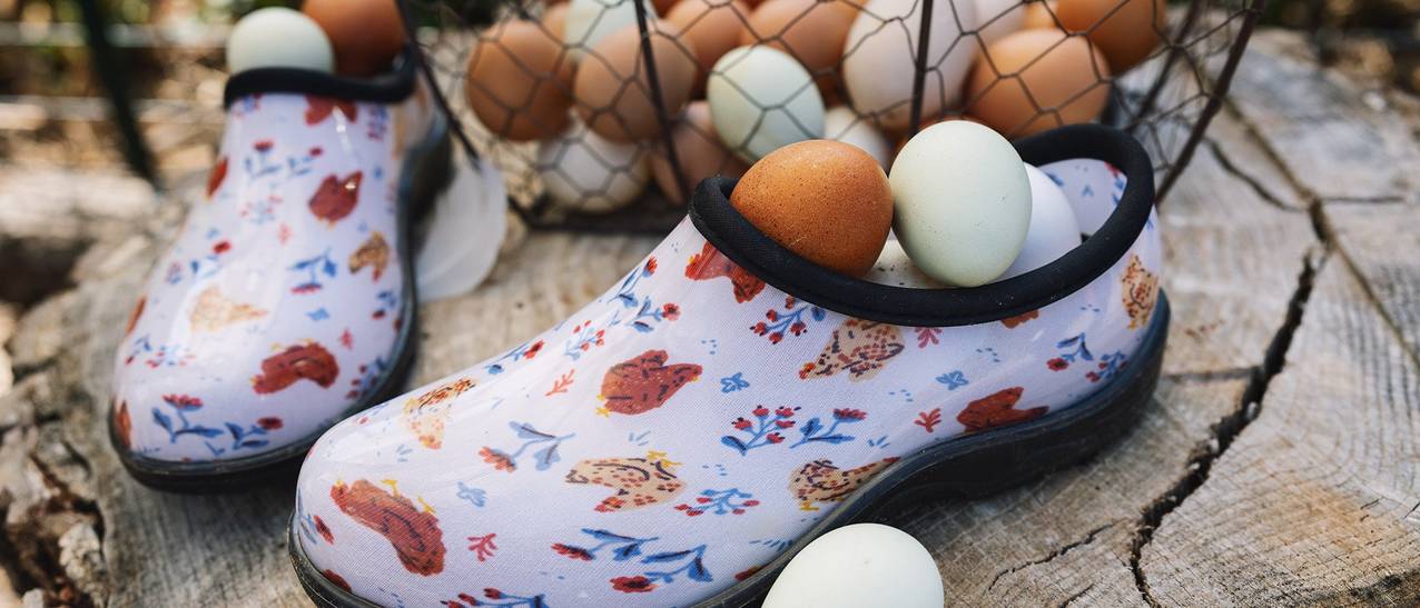 A pair of garden clogs with a chicken print sit next to a basket of eggs