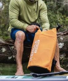 man unpacking an orange duffle bag; sitting on a log with a paddle board