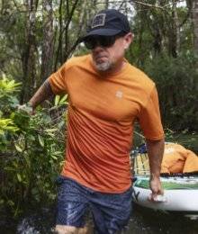 man wearing orange swim shirt walking through a stream holding a paddle board
