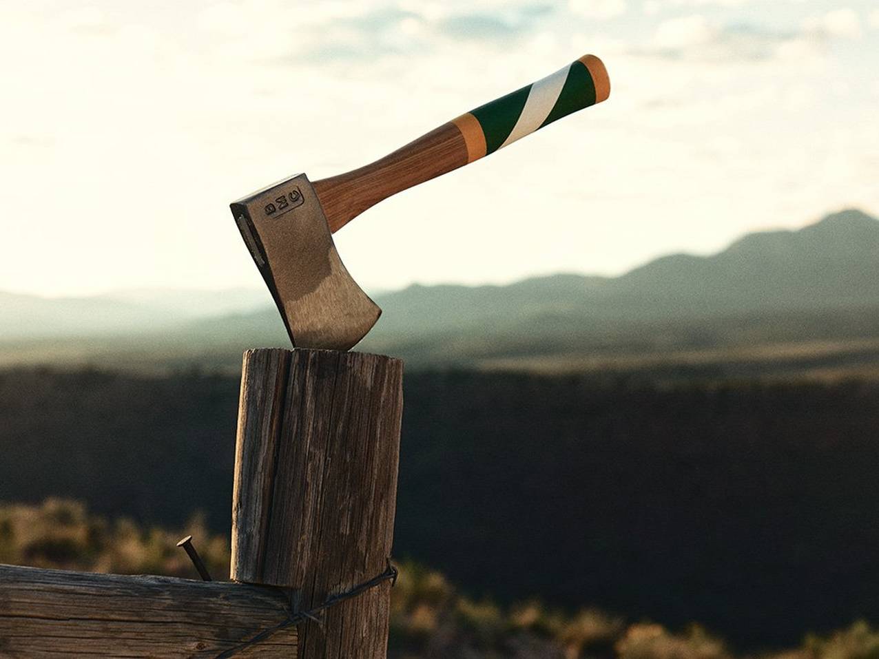 A Best Made hatchet with a colorful handle sticks into a wooden fence post in front of a mountain scene.