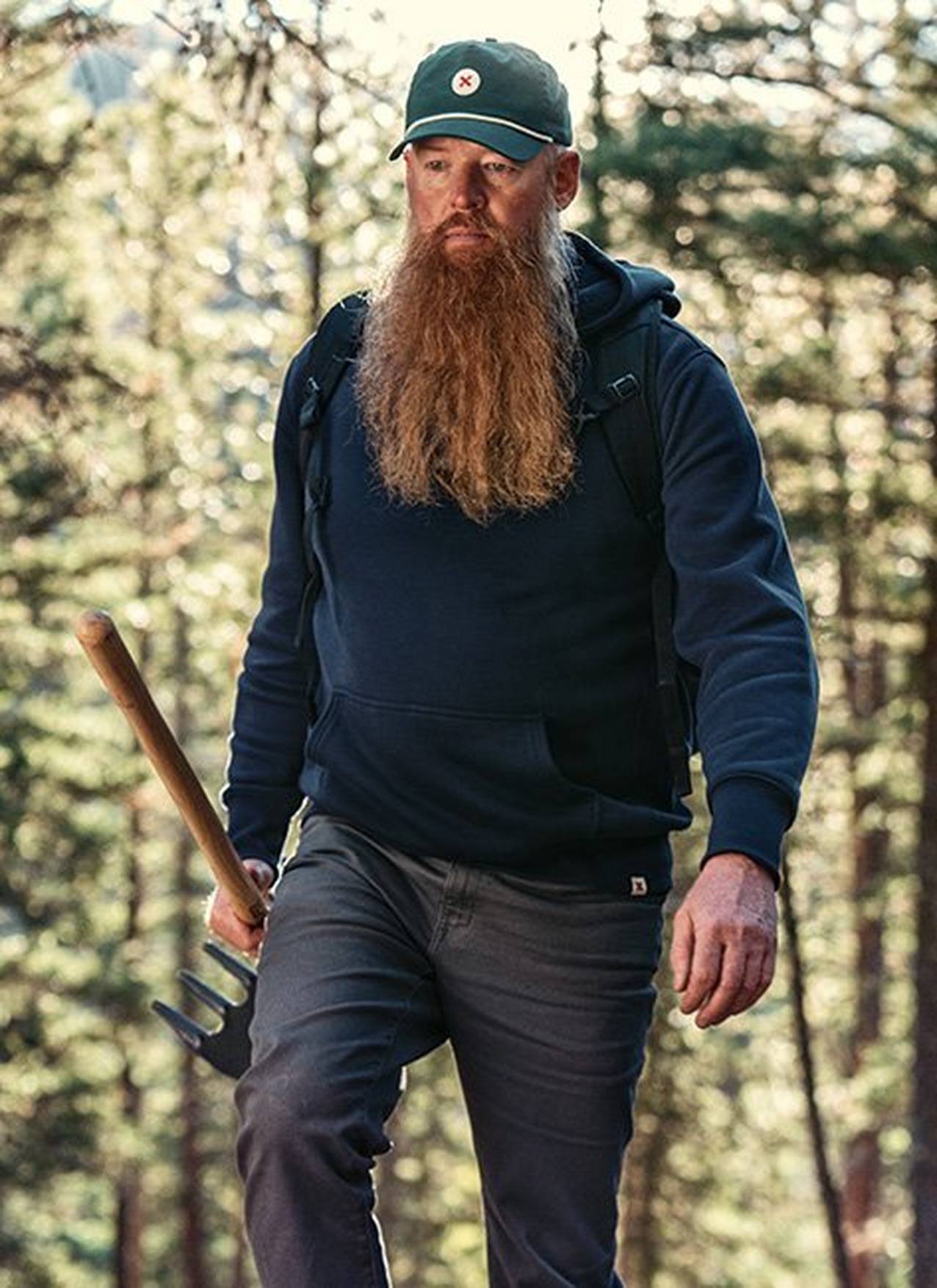 Tom, wearing a green Best Made hat and blue headed sweatshirt, walks through the woods with a rake.