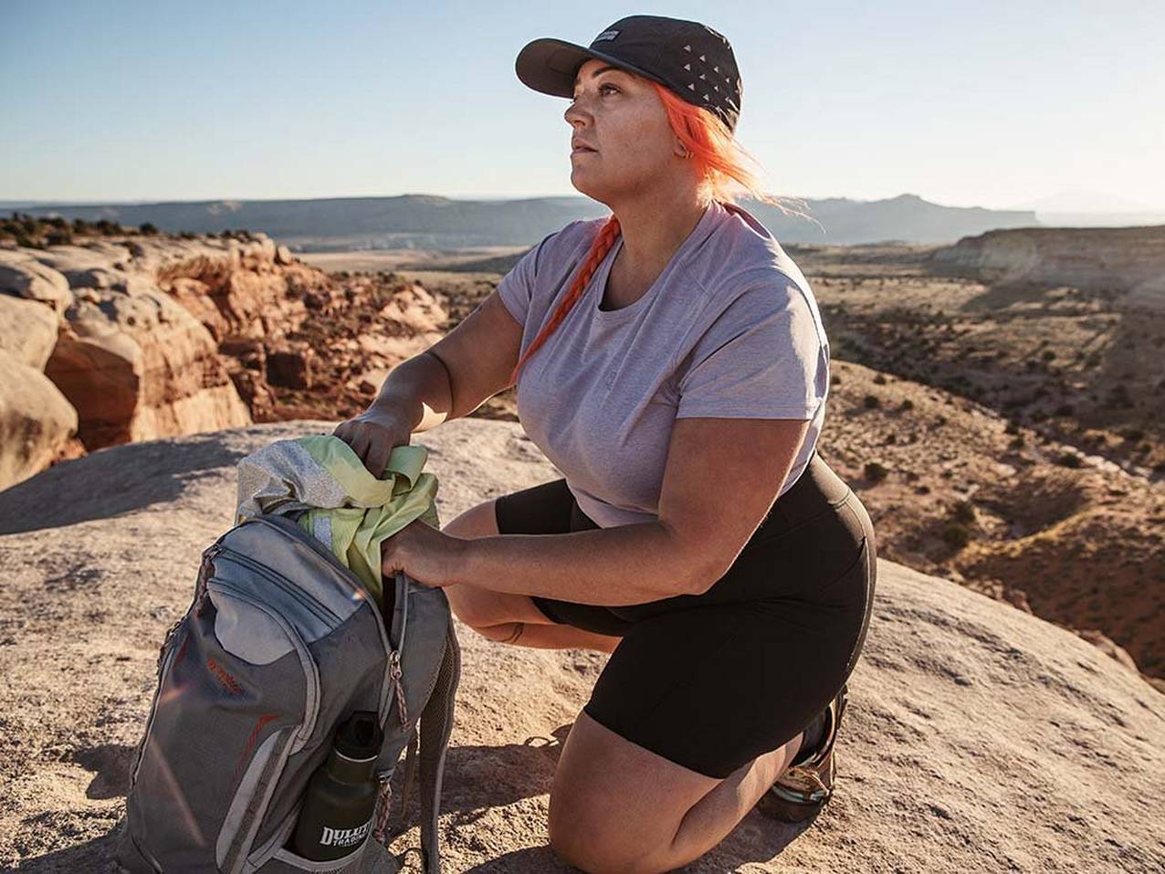 a woman crouching on top of a natural rock formation reaches into her backpack for her jacket