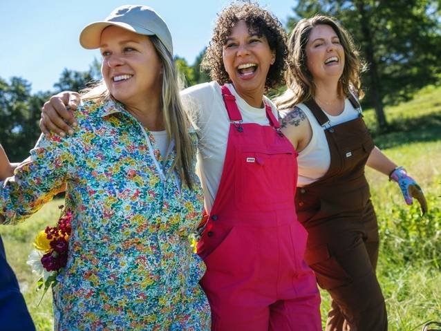 Three women wearing overalls linking arms, smiling.