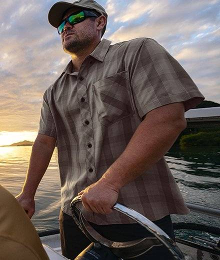A man in a gray plaid shirt drives a boat from the steering wheel.