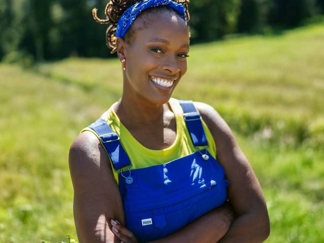 Woman wearing blue overalls, crossing her arms and smiling.