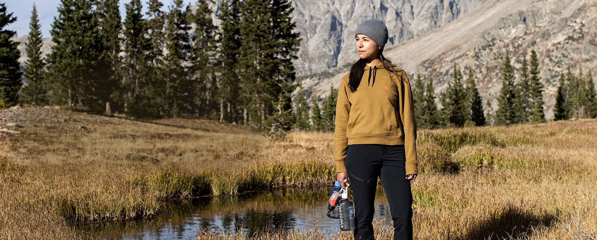 Woman stands by small pond in gold sweatshirt and black pants