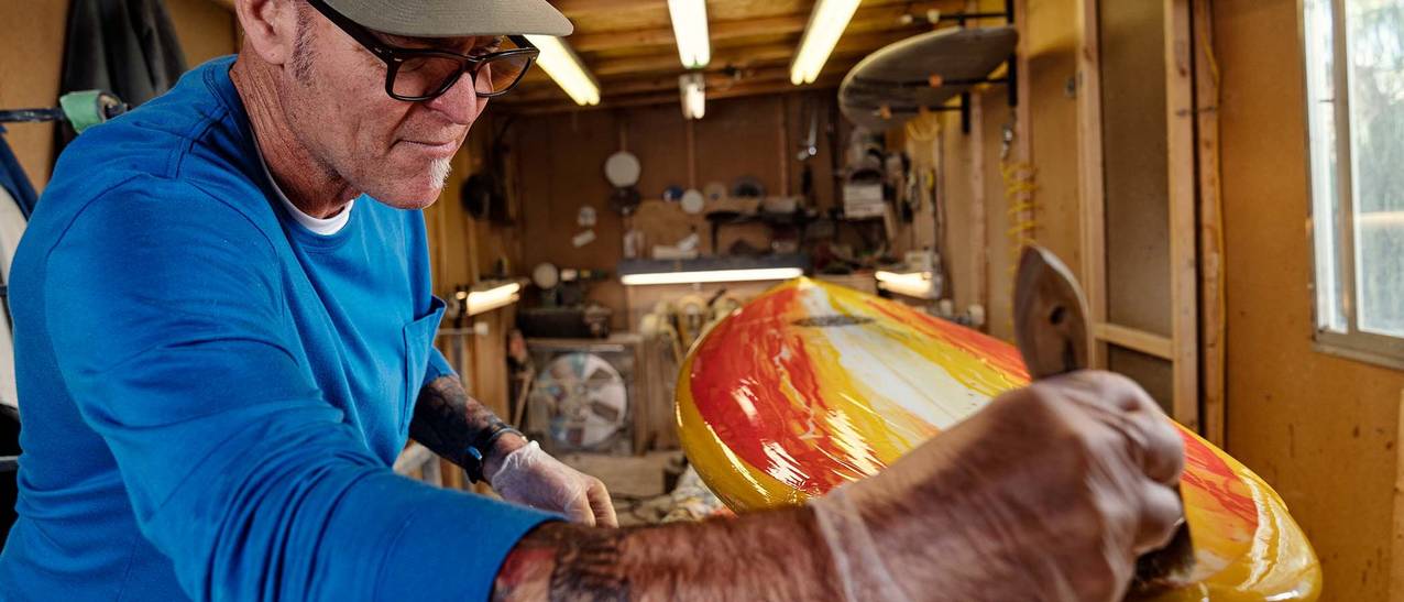 man in a blue shirt and a tan hat coats a surf board