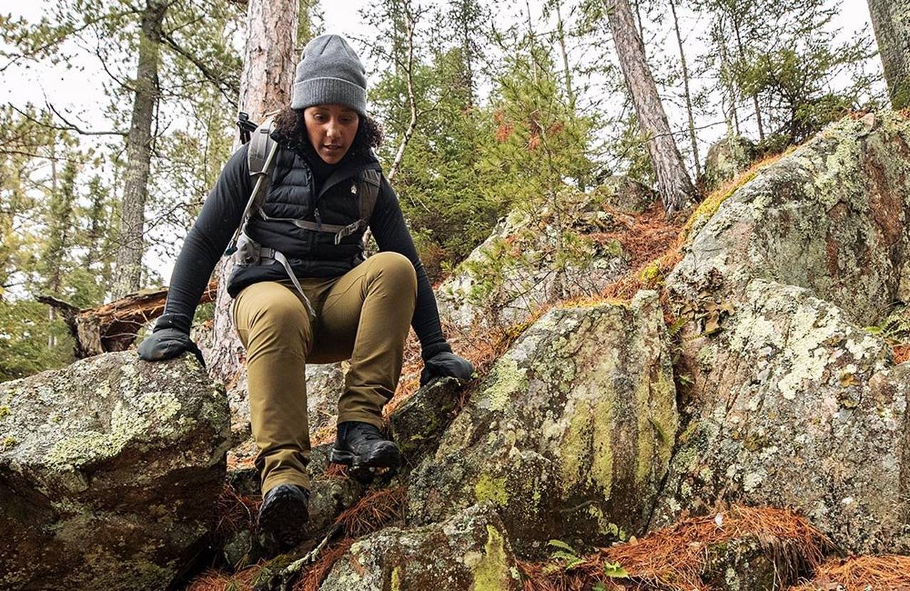 athletic woman in full hiking gear, scrambling down a rocky slope with pine trees behind her