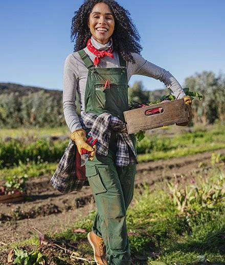 A woman in green overalls carrying a box of veggies