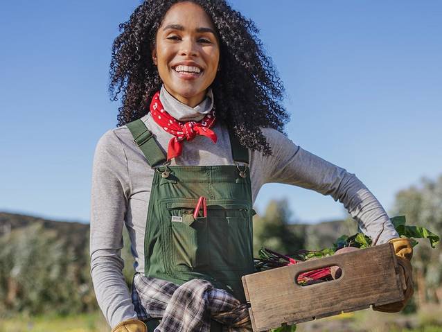 A woman in green overalls stands smiling at the camera with a box of vegetables under one arm