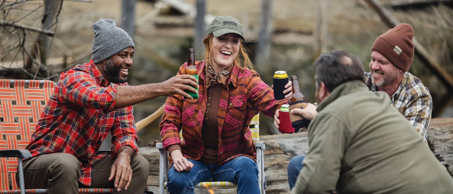 four flannel-clad friends in vintage camp chairs, raise their koozied beers in a toast above their campfire