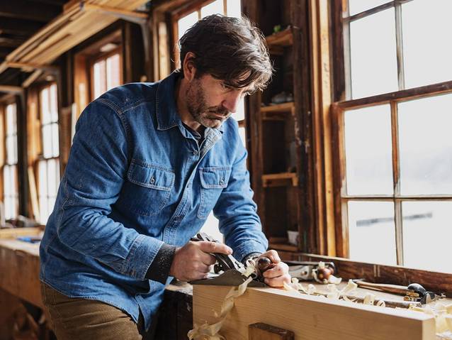 Man wearing a denim collared shirt, planing wood in his boat-building workshop