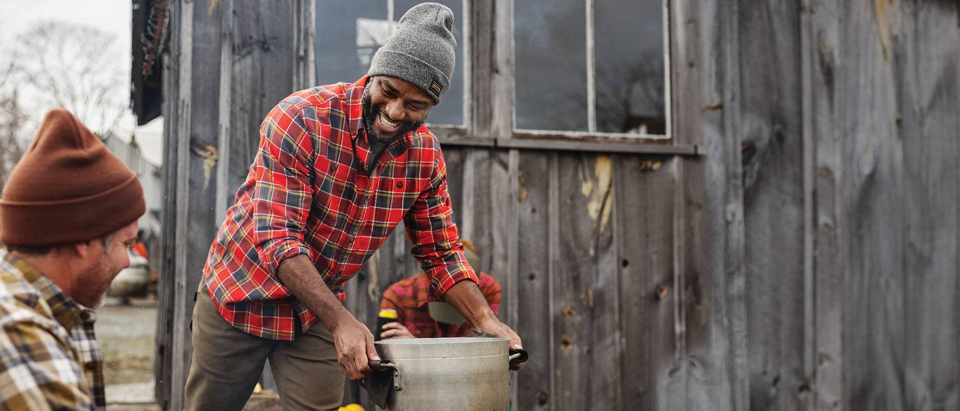 a smiling man in a red flannel shirt carries a stock pot full of oysters to serve his friends outside a fishing shack