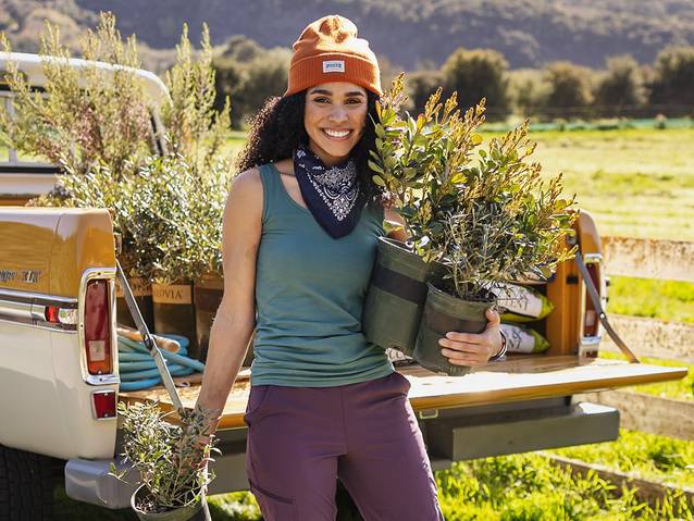 smiling woman wearing a no yank tank, carries potted plants from the tailgate of her truck