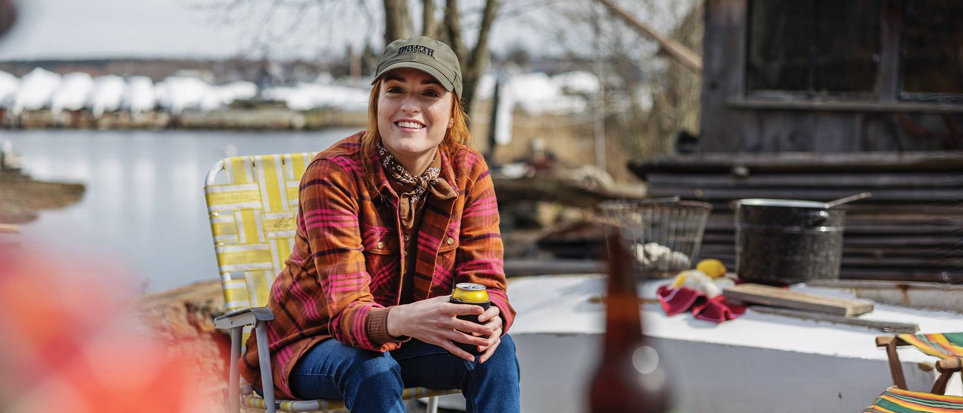 a woman wearing a free swingin' flannel shirt, enjoys a beer with friends in vintage camp chairs near a body of water