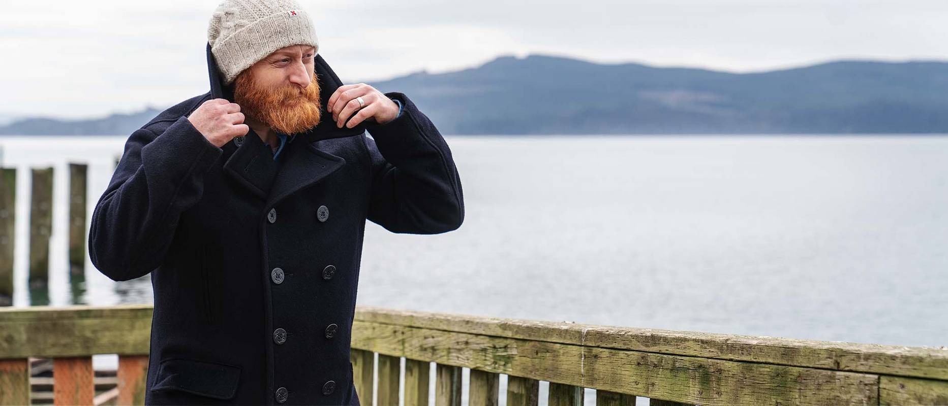 Man with red beard stands on a dock in a pea coat and winter hat