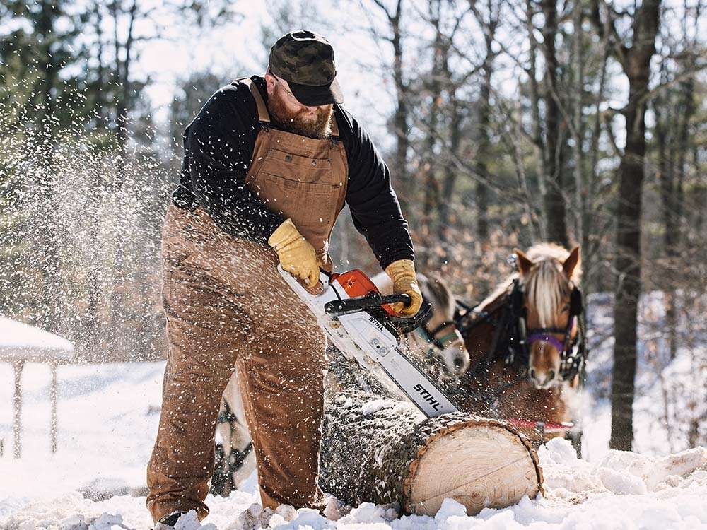 A man in fire hose (r) overalls cuts a log with a chainsaw out in the snow as a horse looks on.