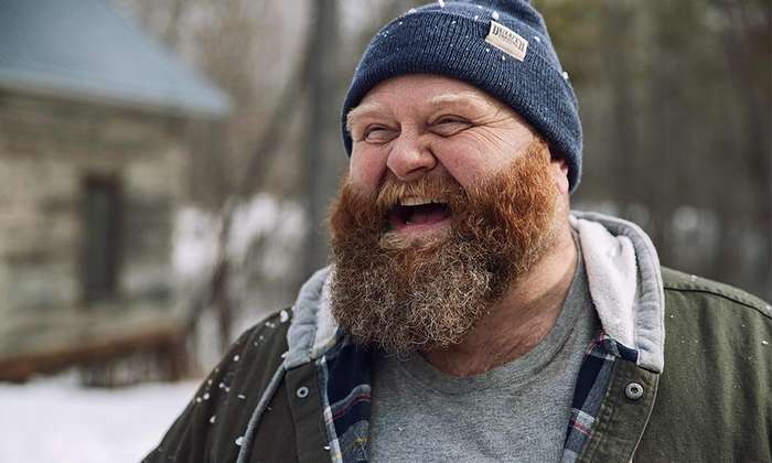 A happy man with a beard stands outside laughing with a blue beanie and hooded jacket