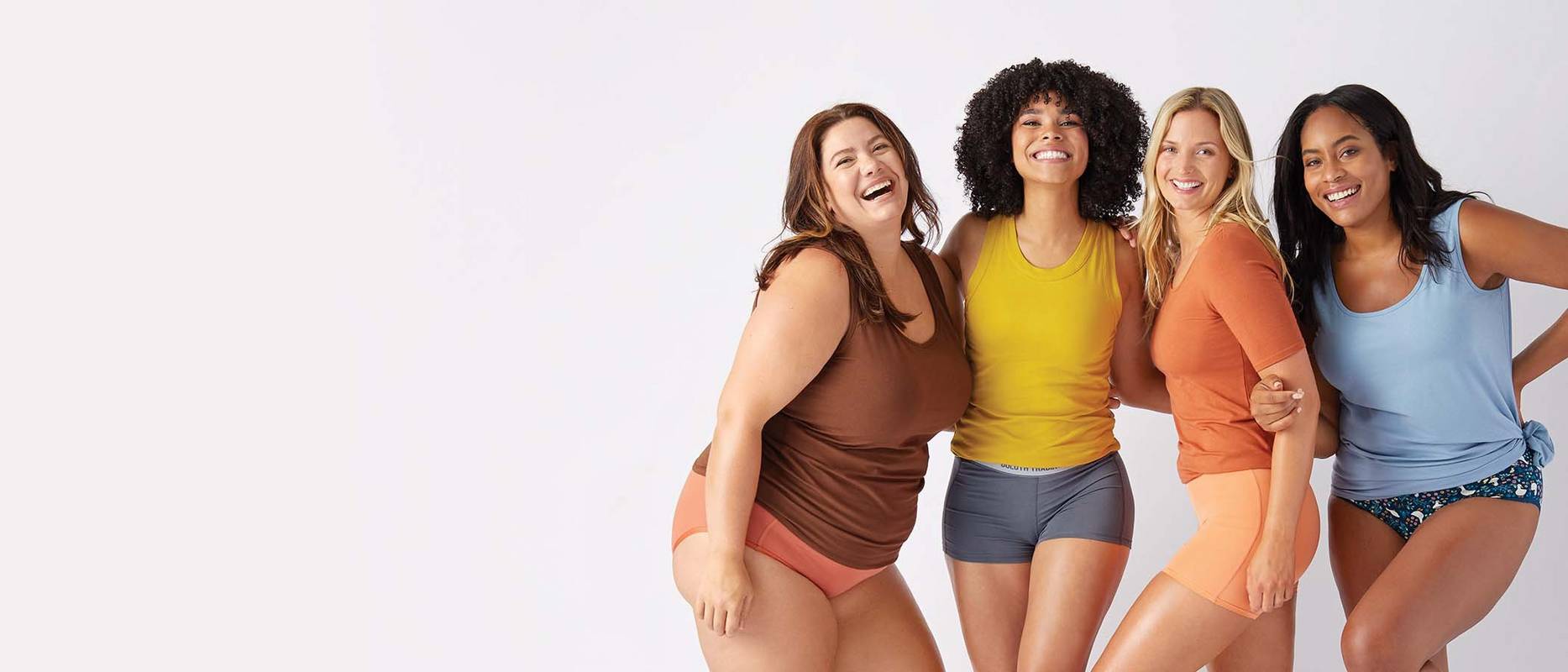 four women standing in their underwear and laughing together against a light studio background