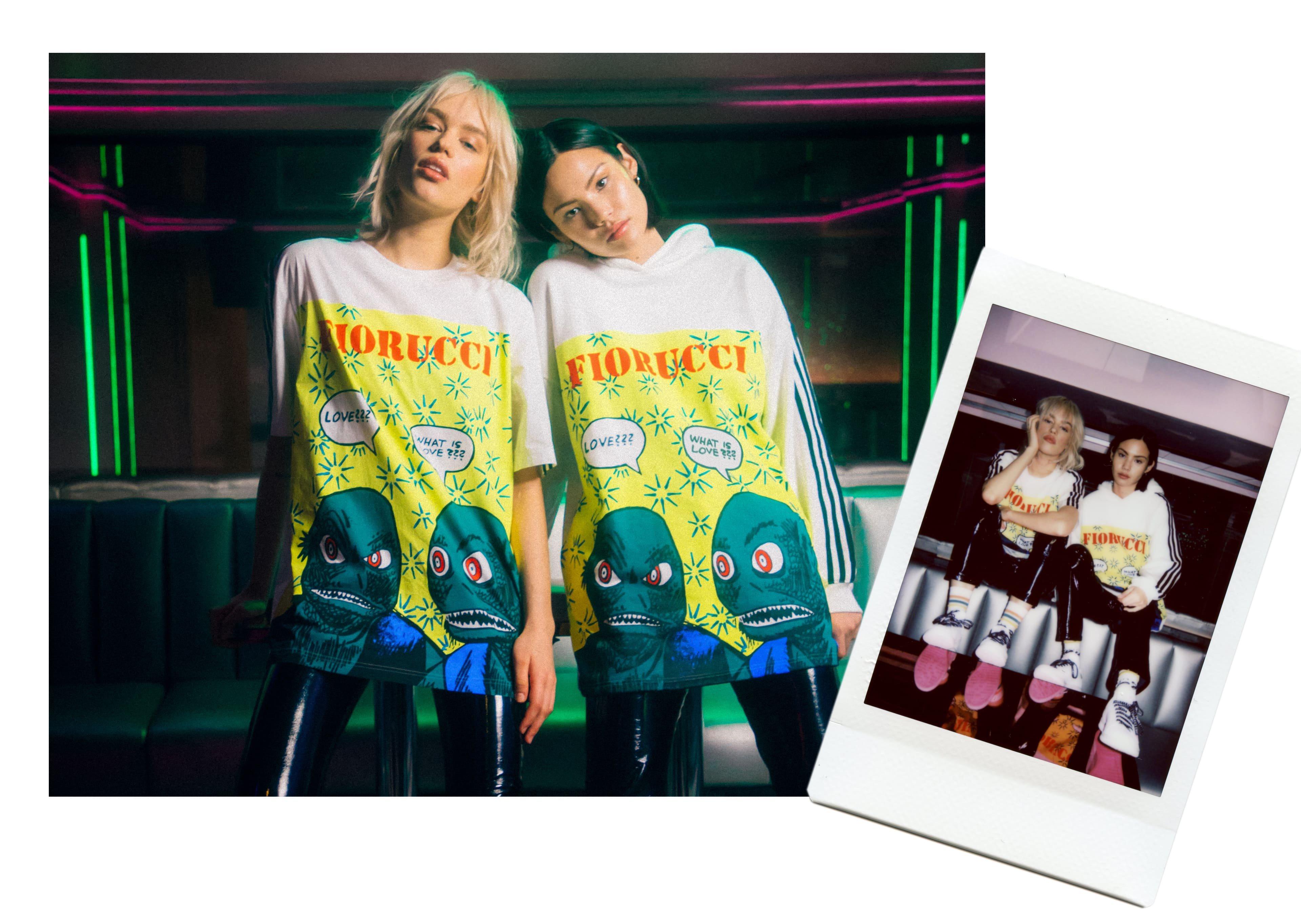 Models wearing the Adidas x Fiorucci Graphic Print Oversized T-Shirt in white and the Adidas x Fiorucci Graphic Archive Print Hoodie in white.