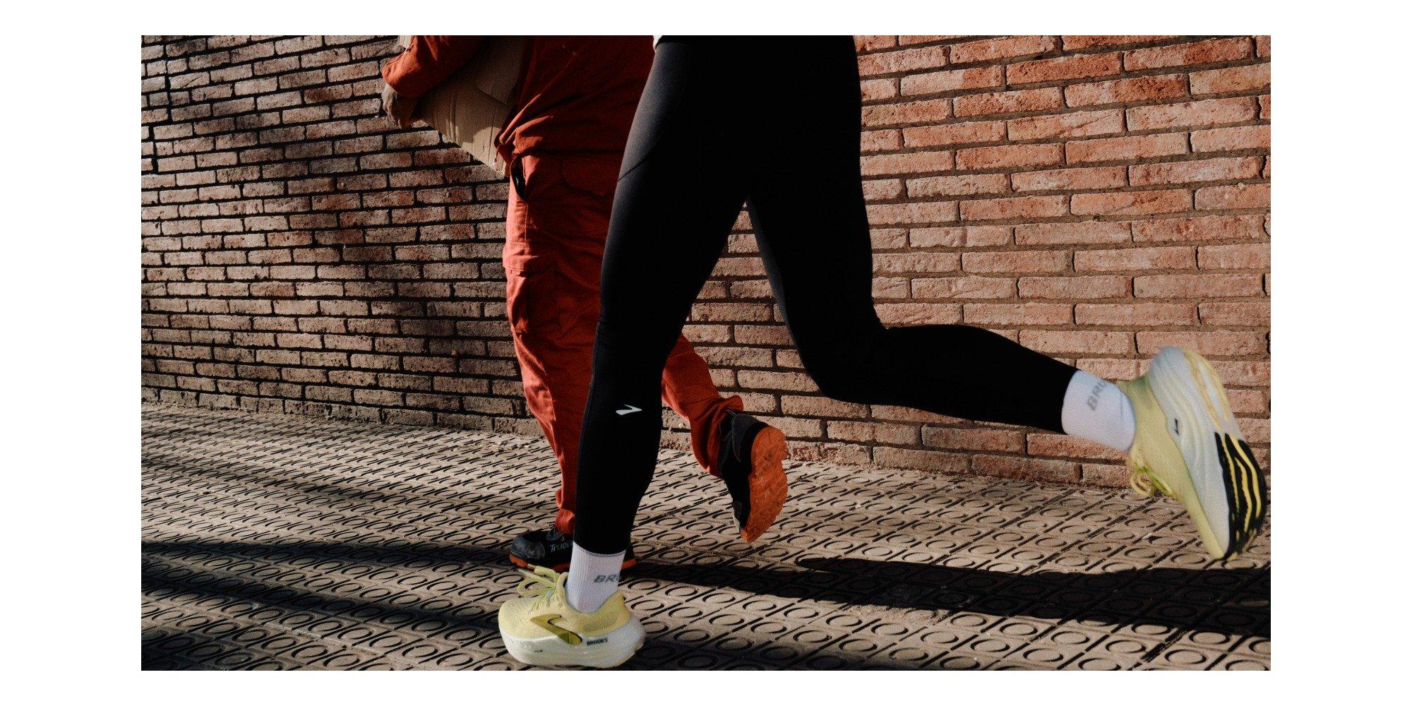 Two people, one running and one walking next to a brick wall on a pavement