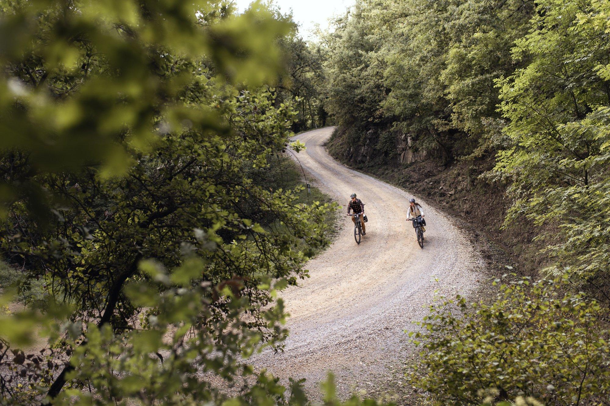two people riding bikes down a dirt road surrounded by trees
