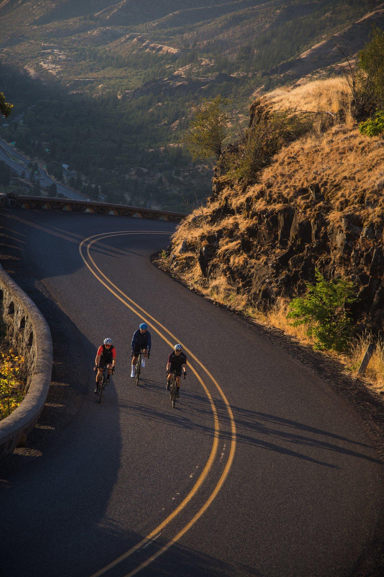 three bicyclists riding down a winding road with a mountain in the background