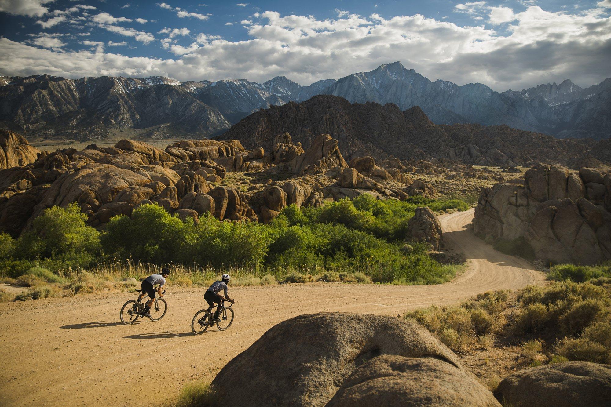 gravel cyclists riding on dirt road with mountains in background