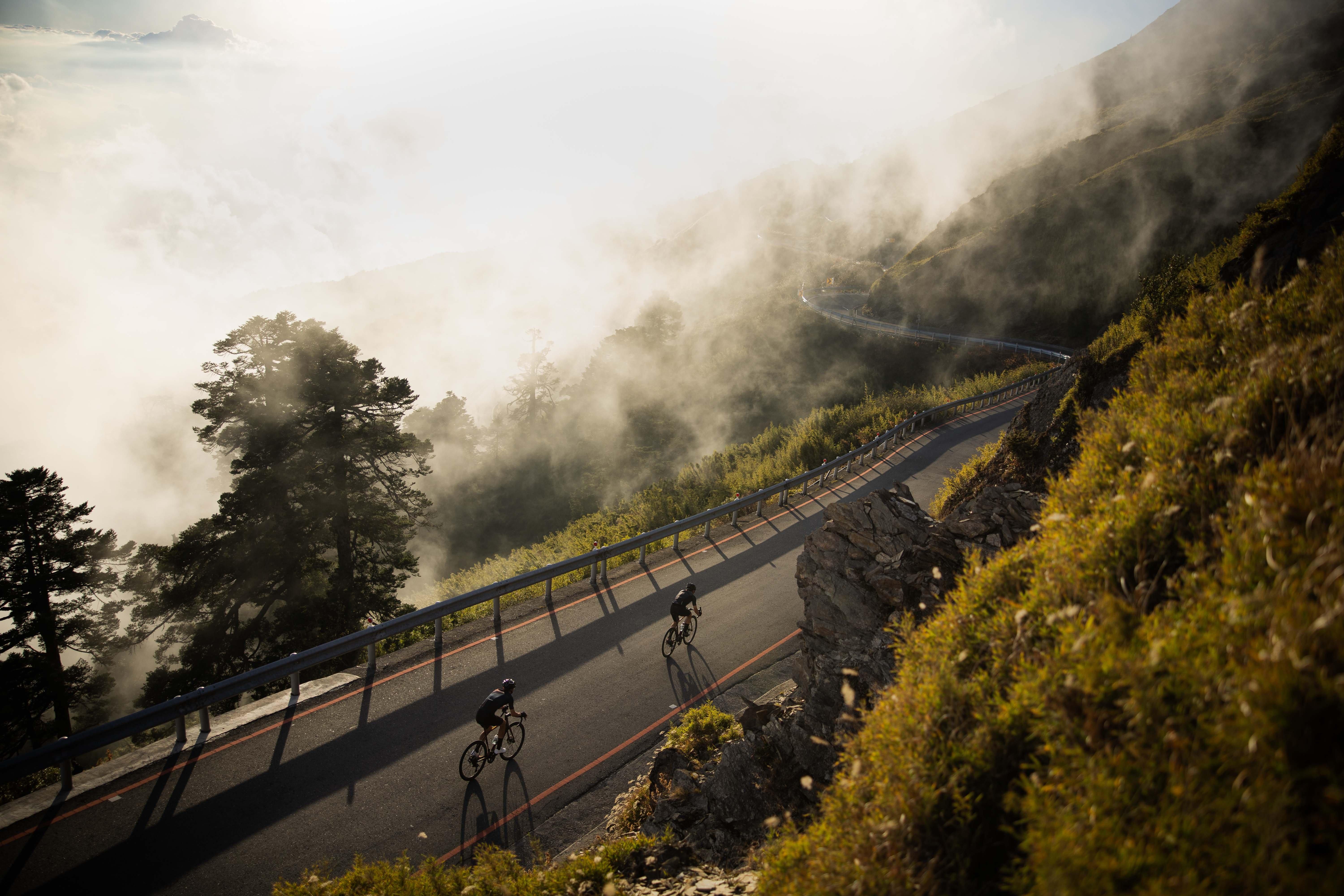 two bicyclists ride down a mountain road in the fog