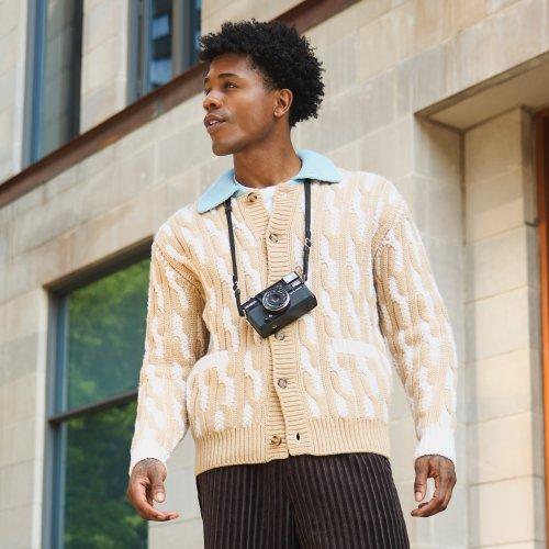 Homme portant un cardigan beige à motifs avec un appareil photo au cou, debout à l’extérieur