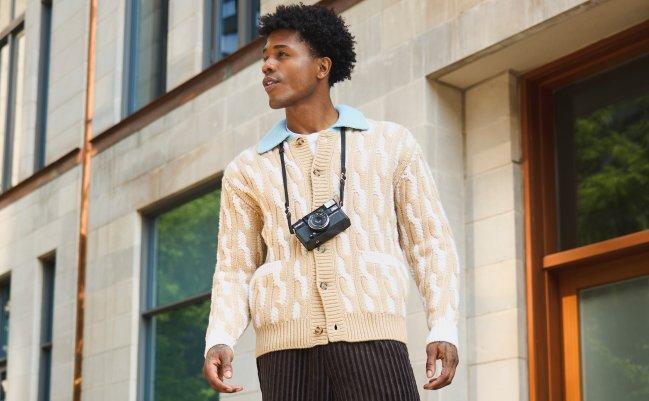Homme portant un cardigan beige à motifs avec un appareil photo au cou, debout à l’extérieur