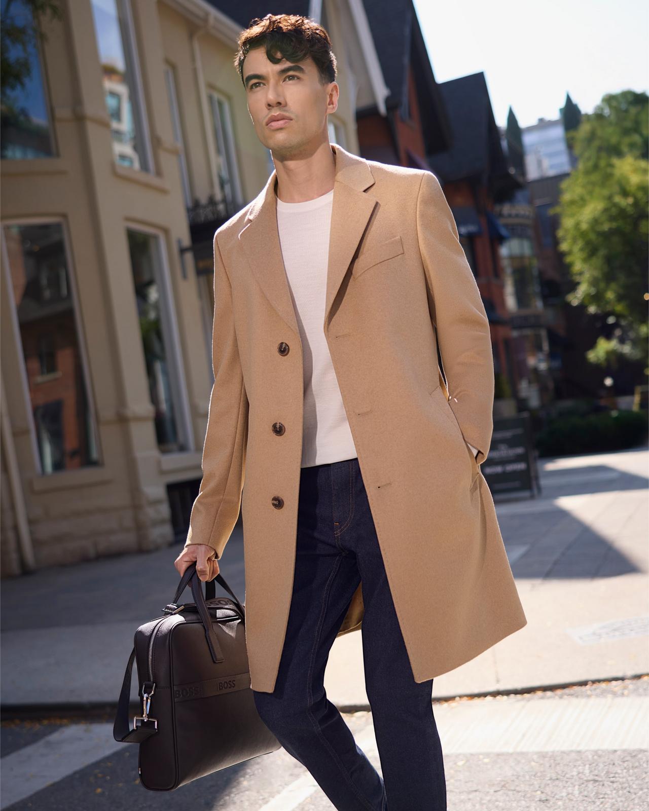 A male model wearing a tan colored BOSS wool-cashmere coat, white T-shirt, and black jeans paired with a brown leather briefcase