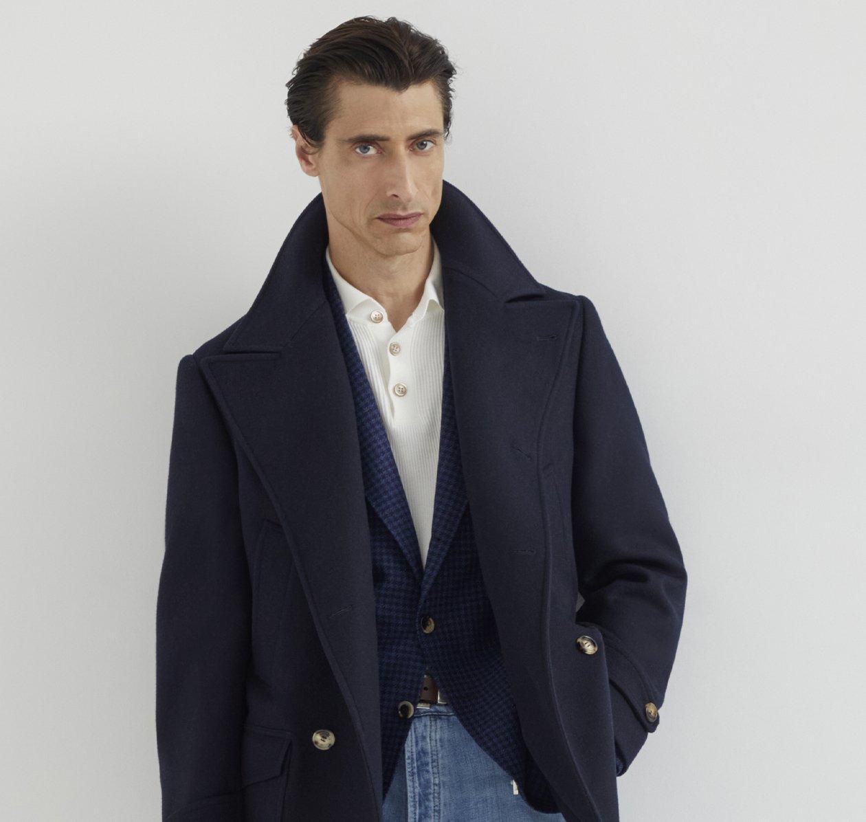 A man wearing a navy Brunello Cucinelli coat over a blue blazer, paired with a white shirt and blue jeans