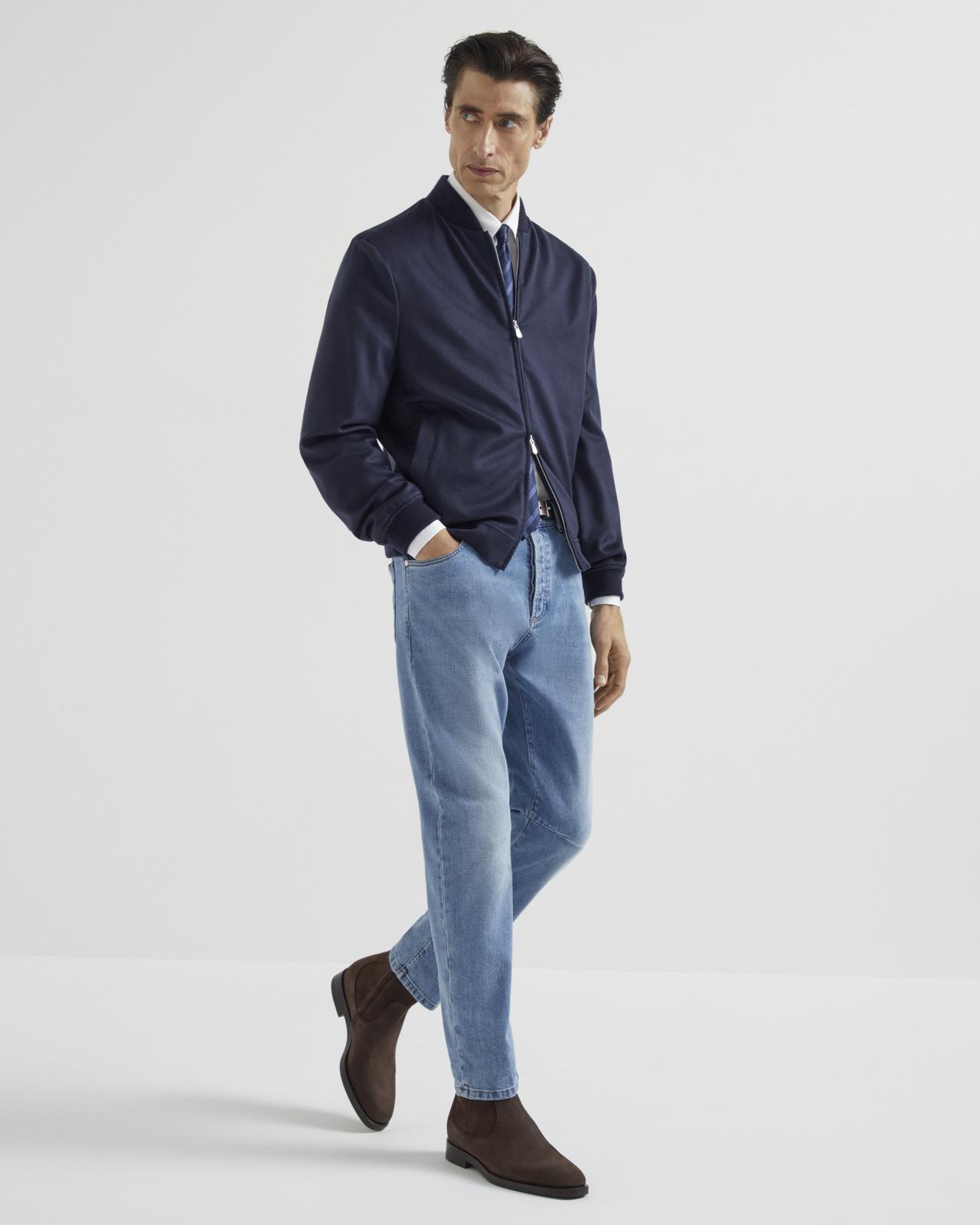 A man wearing a navy Brunello Cucinelli bomber jacket with a white shirt, tie, blue jeans, and brown shoes
