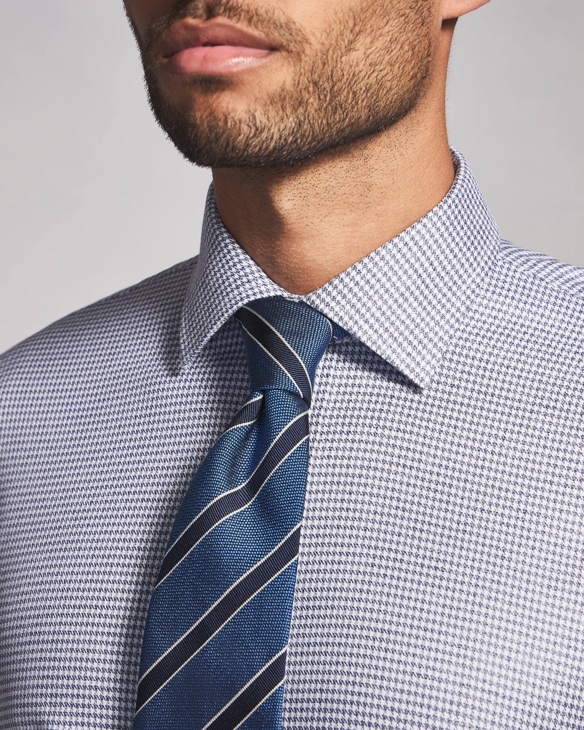 A close up of a model wearing a blue tie with black stripes, paired with a houndstooth casual shirt